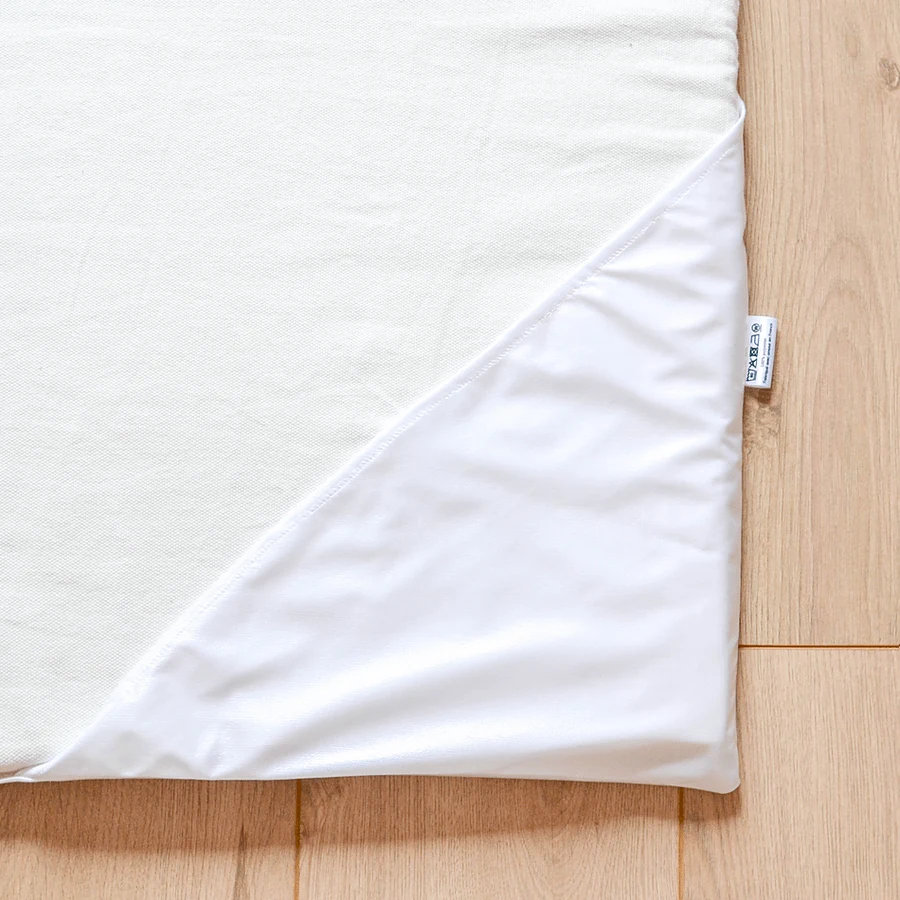 Le coin d'un Tapis Magique dans son alèse écrue, l'insert imperméable blanc glissé par-dessus en triangle, son étiquette cousue sur le bord