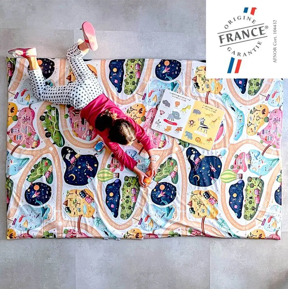 Une petite fille allongée sur un grand tapis imprimé de routes et de maisons, avec en médaillon le logo Origine France Garantie et la mention AFNOR certificat 104432