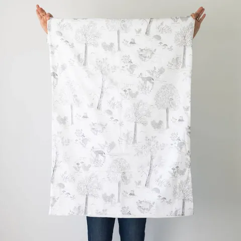 Couverture doux sous-bois dépliée, tenue à bout de bras devant un mur blanc