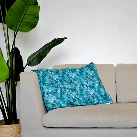 Oreiller housse bleu canard à feuillages posé sur un canapé beige, grande plante verte