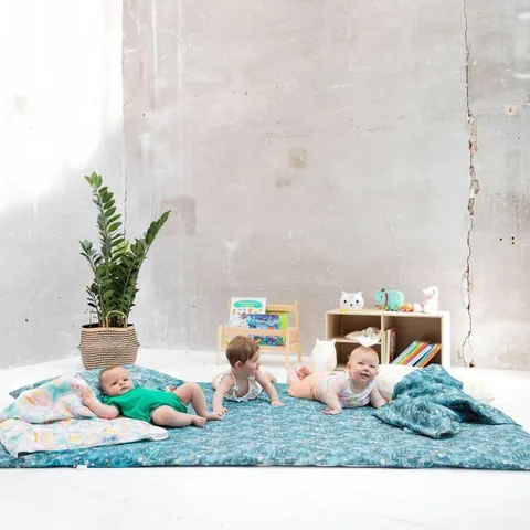 Trois bébés sur le grand tapis bleu canard à feuillages, couverture et oreiller à gauche, plan large