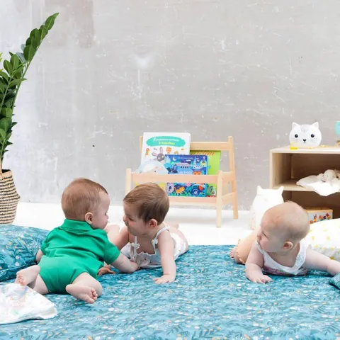 Trois bébés à quatre pattes sur le tapis bleu canard à feuillages, bibliothèque basse, plan rapproché