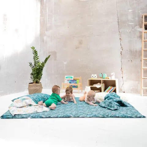 Trois bébés sur le grand tapis bleu canard à feuillages, plante, bibliothèque et échelle, plan large