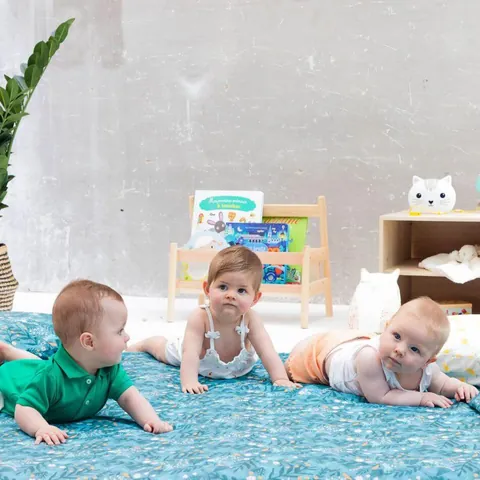 Trois bébés sur le tapis bleu canard à feuillages, l'un en vert regarde l'objectif, plan rapproché