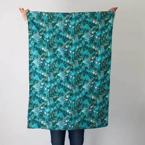 Couverture forêt magique dépliée, tenue à bout de bras devant un mur blanc