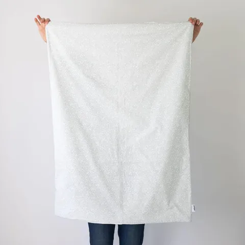Couverture vert de gris à petites fleurs dépliée, tenue à bout de bras devant un mur blanc