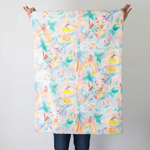 Couverture jungle aquarelle pastel dépliée, tenue à bout de bras devant un mur blanc