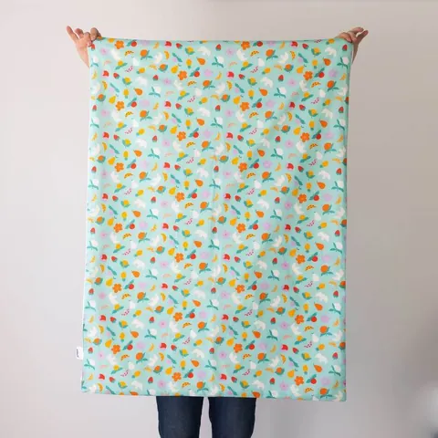 Couverture vert d'eau à fruits et oiseaux dépliée, tenue à bout de bras devant un mur blanc