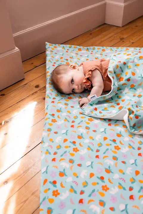 Bébé allongé sur le côté, emmitouflé dans la couverture Pique-nique, tapis assorti, soleil sur le parquet