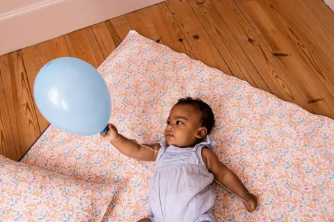 Bébé qui tient un ballon bleu, allongé sur le tapis Poésie, l'oreiller Poésie au premier plan