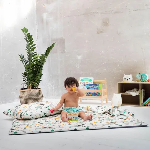 Bébé assis sur le tapis savane avec l'oreiller et la couverture assortis, plante et bibliothèque basse