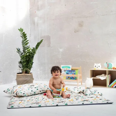 Bébé souriant assis sur le tapis savane, oreiller et couverture assortis, plante et bibliothèque basse