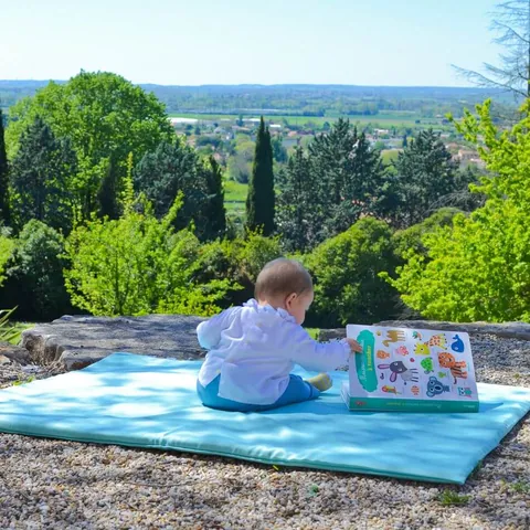 Bébé assis de profil sur le tapis Baroudeur feuilletant un grand livre, vallée en fond