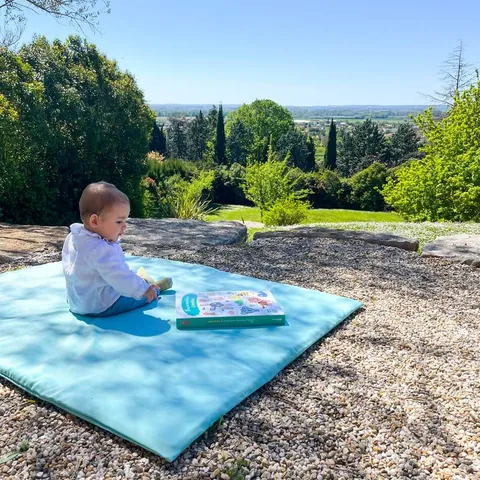 Bébé assis avec un livre sur le Tapis Magique Le baroudeur, dans un jardin face à la vallée