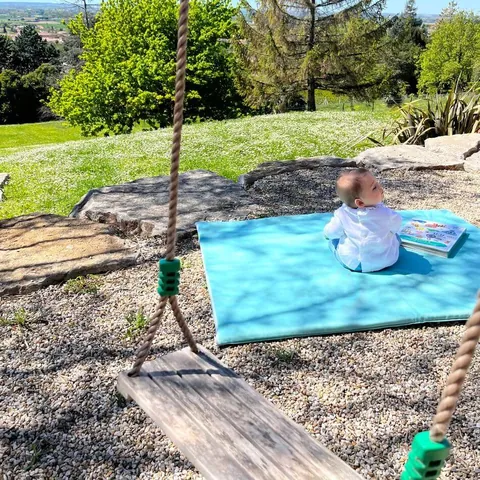 Balançoire en bois au premier plan, bébé de dos sur le tapis Baroudeur dans le jardin