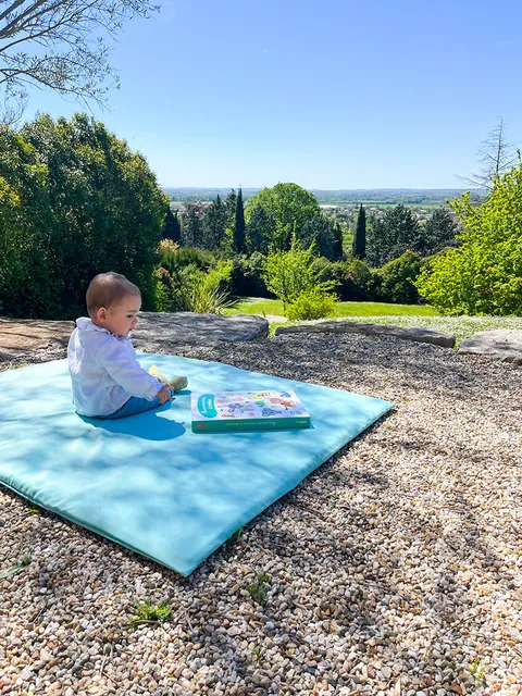 Bébé assis sur le tapis Baroudeur avec un livre, jardin en surplomb et ciel bleu, cadrage vertical