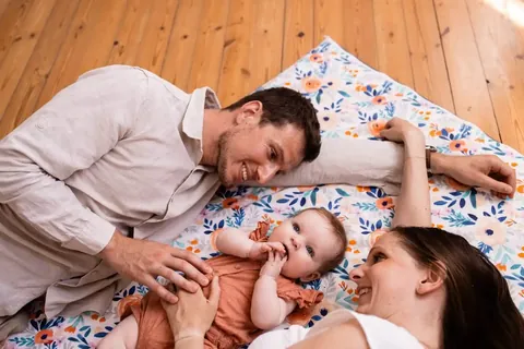Père et mère allongés autour d'un bébé sur le tapis blanc à fleurs orange et bleues, parquet