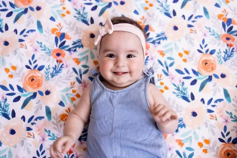 Bébé souriant au bandeau rose allongé sur le tapis blanc à fleurs orange et bleues, vue de dessus