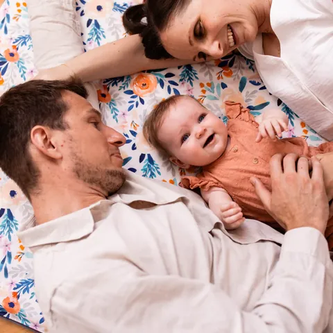 Père et mère allongés tête contre tête autour d'un bébé souriant sur le tapis blanc à fleurs orange et bleues, vue de dessus
