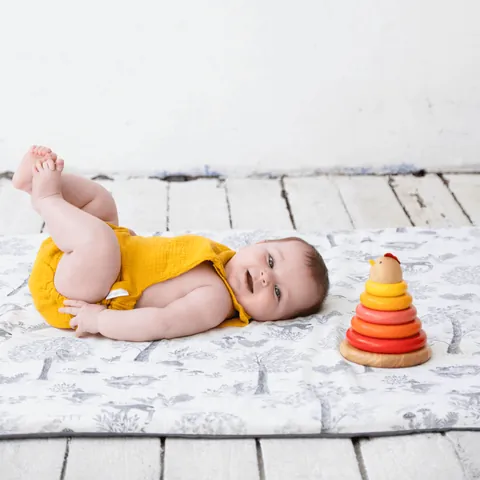 Bébé en barboteuse jaune jambes levées sur le tapis blanc à dessins gris de sous-bois, pyramide d'anneaux, plan rapproché