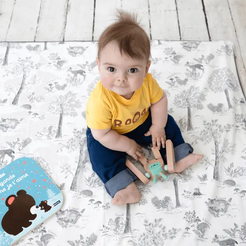 Bébé en tee-shirt jaune assis sur le tapis blanc à dessins gris de sous-bois, jouet en bois, livre, vue plongeante