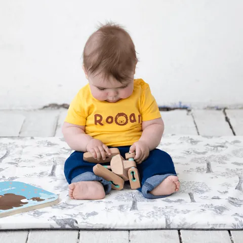 Bébé en t-shirt jaune assis sur le tapis Doux sous-bois, tête baissée sur un jouet en bois, livre à côté