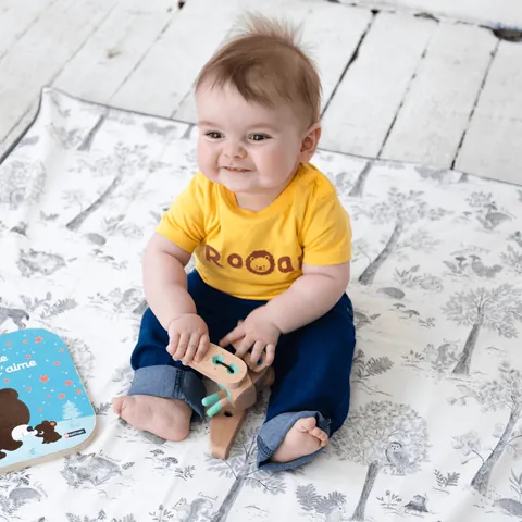 Bébé souriant en tee-shirt jaune assis sur le tapis blanc à dessins gris de sous-bois, jouet en bois, vue plongeante