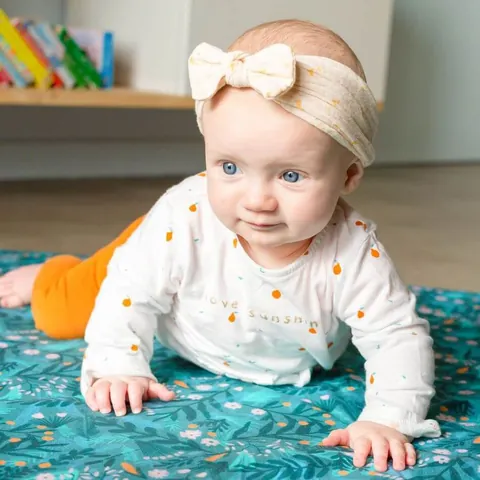 Bébé au bandeau à noeud appuyé sur les bras sur le tapis Forêt magique, étagère de livres en fond