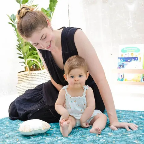 Femme assise et bébé assis contre elle sur le tapis vert d'eau à feuillages, plante et étagère en fond