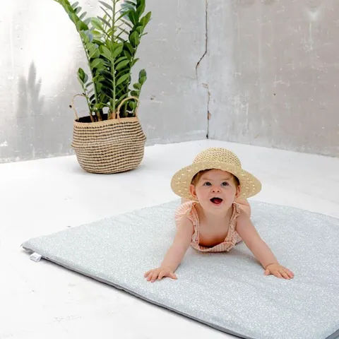Bébé au chapeau de paille qui rit, sur le ventre sur le tapis vert de gris à petites fleurs blanches, plante en pot