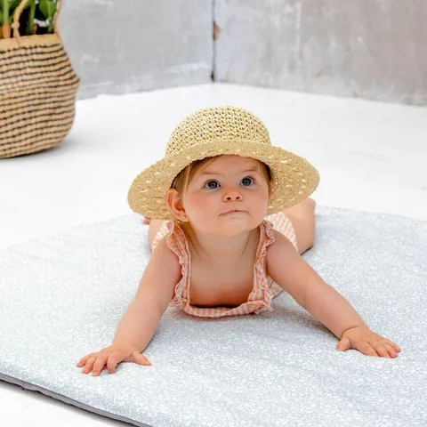 Petite fille au chapeau de paille allongée sur le ventre sur le tapis Gabi, regard vers le haut, plante en fond