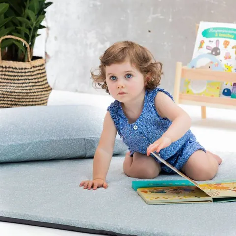 Petite fille à quatre pattes qui feuillette un livre sur le tapis vert de gris à petites fleurs blanches, coussin assorti