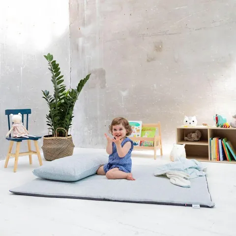 Petite fille assise sur le tapis Gabi avec coussin, chaise, plante, présentoir de livres et étagère