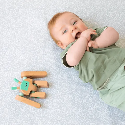 Bébé qui suce ses doigts, allongé sur le tapis vert de gris à petites fleurs blanches, jouet en bois, plan rapproché