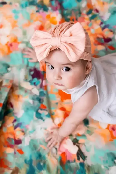 Bébé au gros noeud rose qui lève les yeux, sur le ventre sur le tapis à fleurs peintes turquoise et orange, vue de dessus