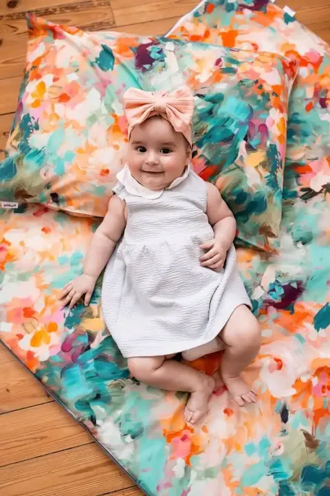 Bébé en robe blanche allongé sur le tapis à fleurs peintes turquoise et orange, coussin assorti, vue plongeante