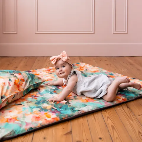 Bébé au noeud rose sur le ventre sur le tapis à fleurs peintes turquoise et orange, coussin assorti, parquet et murs roses moulurés