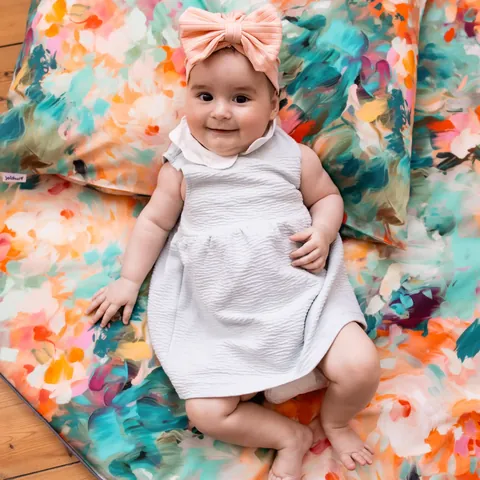 Bébé souriant en robe blanche allongé sur le tapis Jardin, coussin assorti derrière, vue du dessus