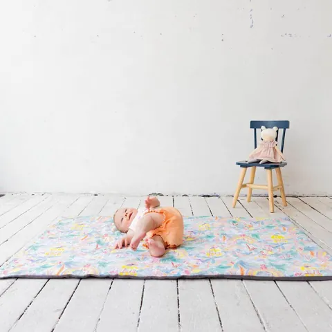 Plan large : bébé allongé sur le tapis Jungle enchantée sur un plancher blanc, petite chaise et peluche au mur