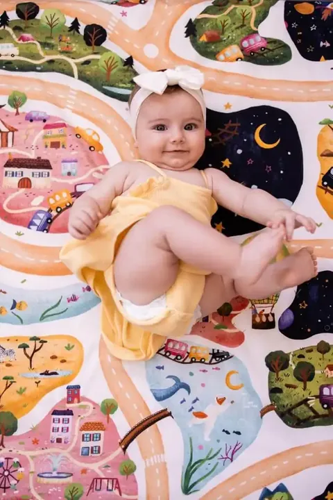 Bébé au bandeau blanc allongé qui attrape son pied sur le tapis Croisée des mondes (routes, villages, ciel étoilé), vue de dessus