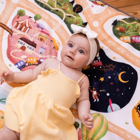 Bébé en robe jaune et bandeau blanc allongé sur l'imprimé À la croisée des mondes, regard vers l'objectif