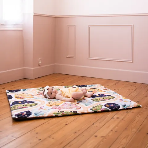 Bébé allongé sur le tapis Croisée des mondes posé sur un parquet, chambre rose moulurée, plan large, tapis entier