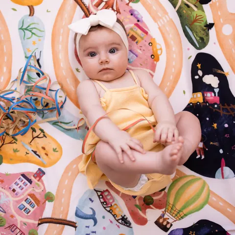 Bébé en robe jaune et bandeau blanc qui tient ses pieds, serpentins, sur l'imprimé À la croisée des mondes, vue du dessus