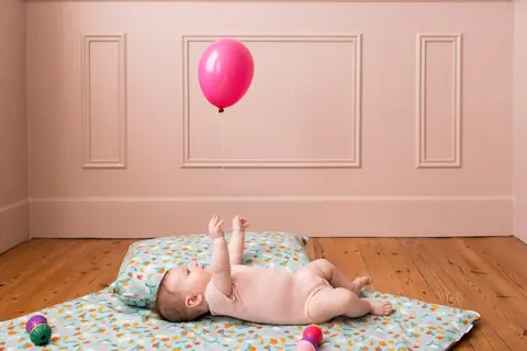 Bébé allongé sur le tapis bleu clair à confettis colorés dans une chambre aux murs roses moulurés, ballon rose
