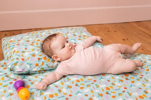 Bébé en body rose allongé sur le tapis bleu clair à confettis colorés, tête sur le coussin, jouet, plan au ras du sol
