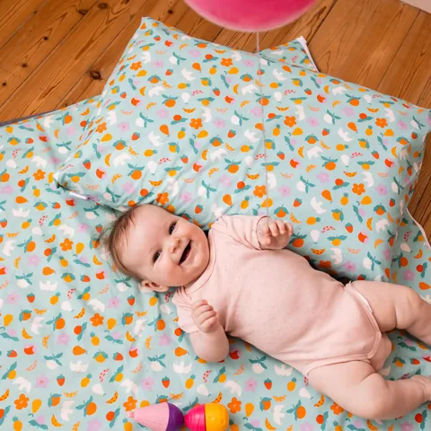 Bébé souriant allongé sur le Tapis Magique Pique-nique estival, la tête sur le coussin assorti