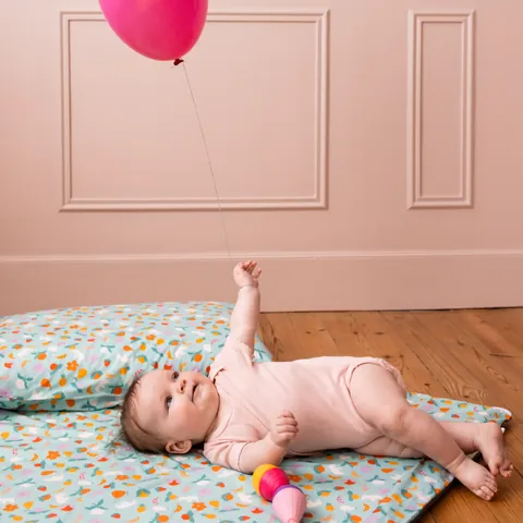 Bébé allongé qui lève le bras vers un ballon rose sur le tapis Pique-nique, coussin assorti, murs roses