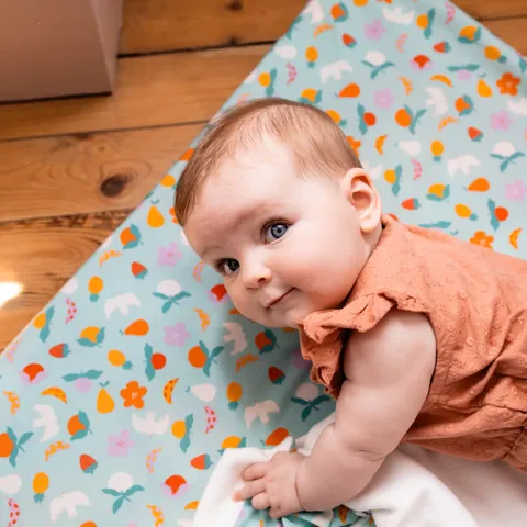 Bébé sur le ventre qui regarde l'objectif, tapis bleu clair à confettis colorés posé sur un parquet, lumière naturelle