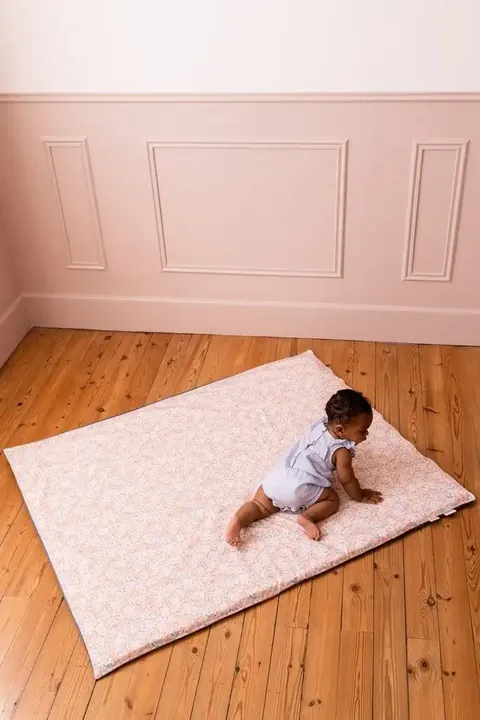 Bébé à quatre pattes sur le tapis rose pâle à petites fleurs posé en biais sur un parquet, chambre aux murs roses