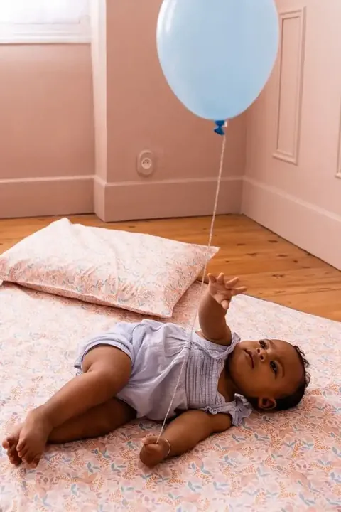Bébé allongé qui tend la main vers un ballon bleu, tapis rose pâle à petites fleurs et coussin assorti, parquet